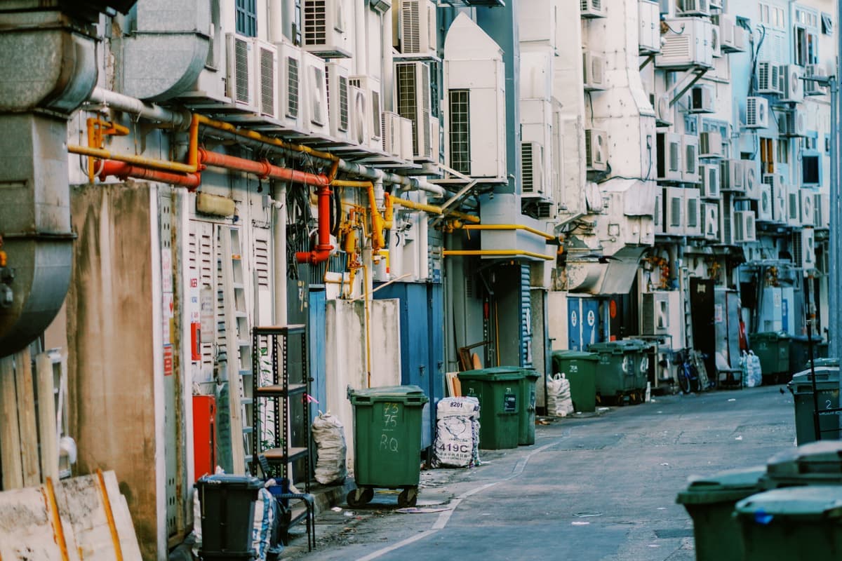 A narrow urban alley lined with air conditioning units and trash bins