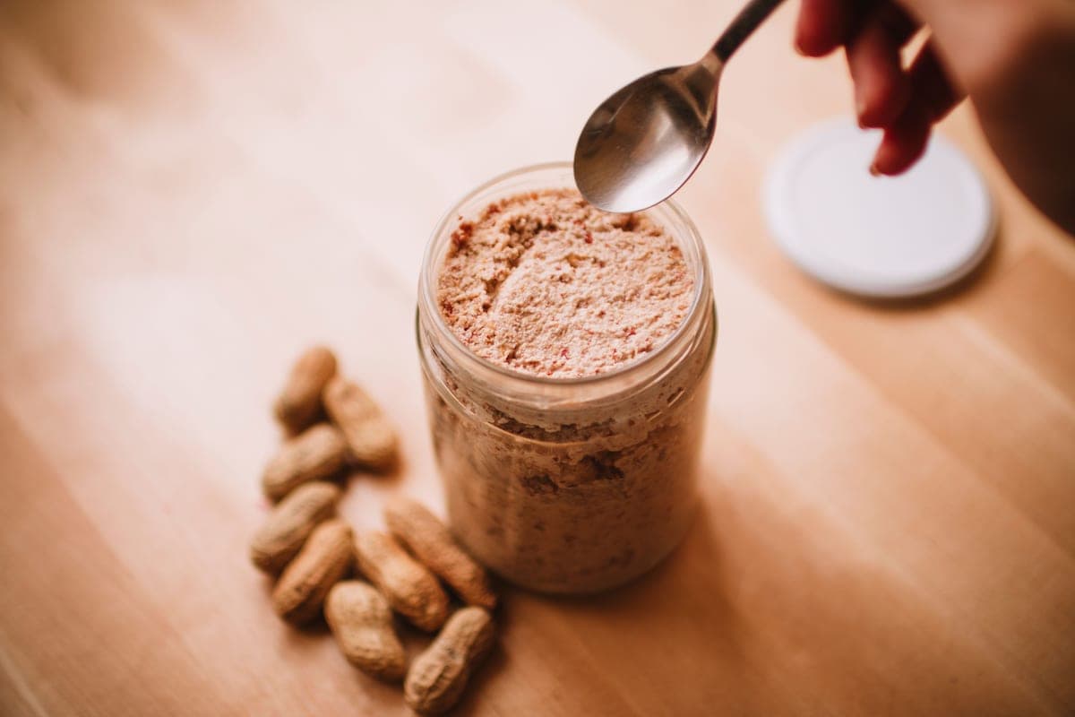 A glass jar of peanut butter on a wooden table with peanuts and a spoon