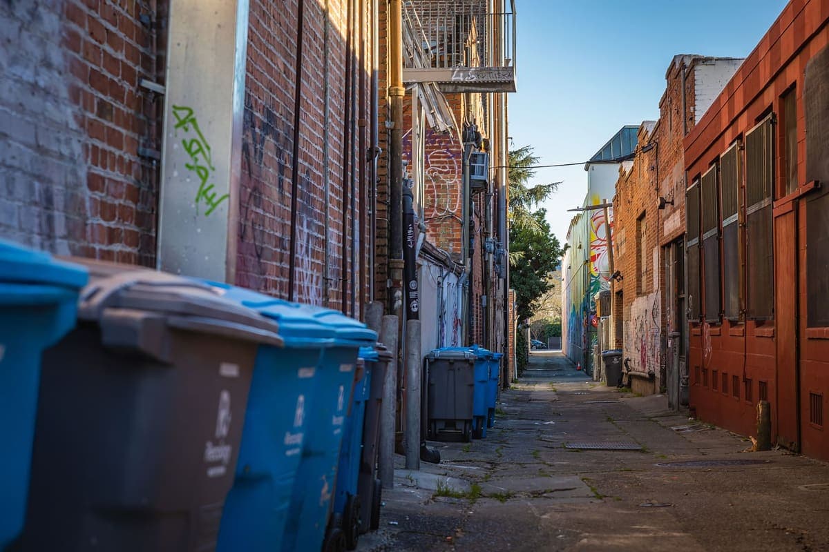 An urban alleyway with blue garbage bins and brick buildings
