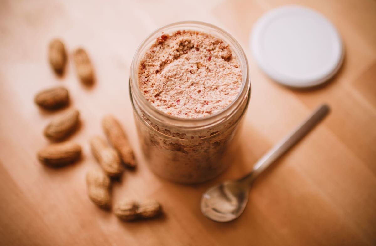 A glass jar of peanut butter with peanuts and a spoon on a wooden table